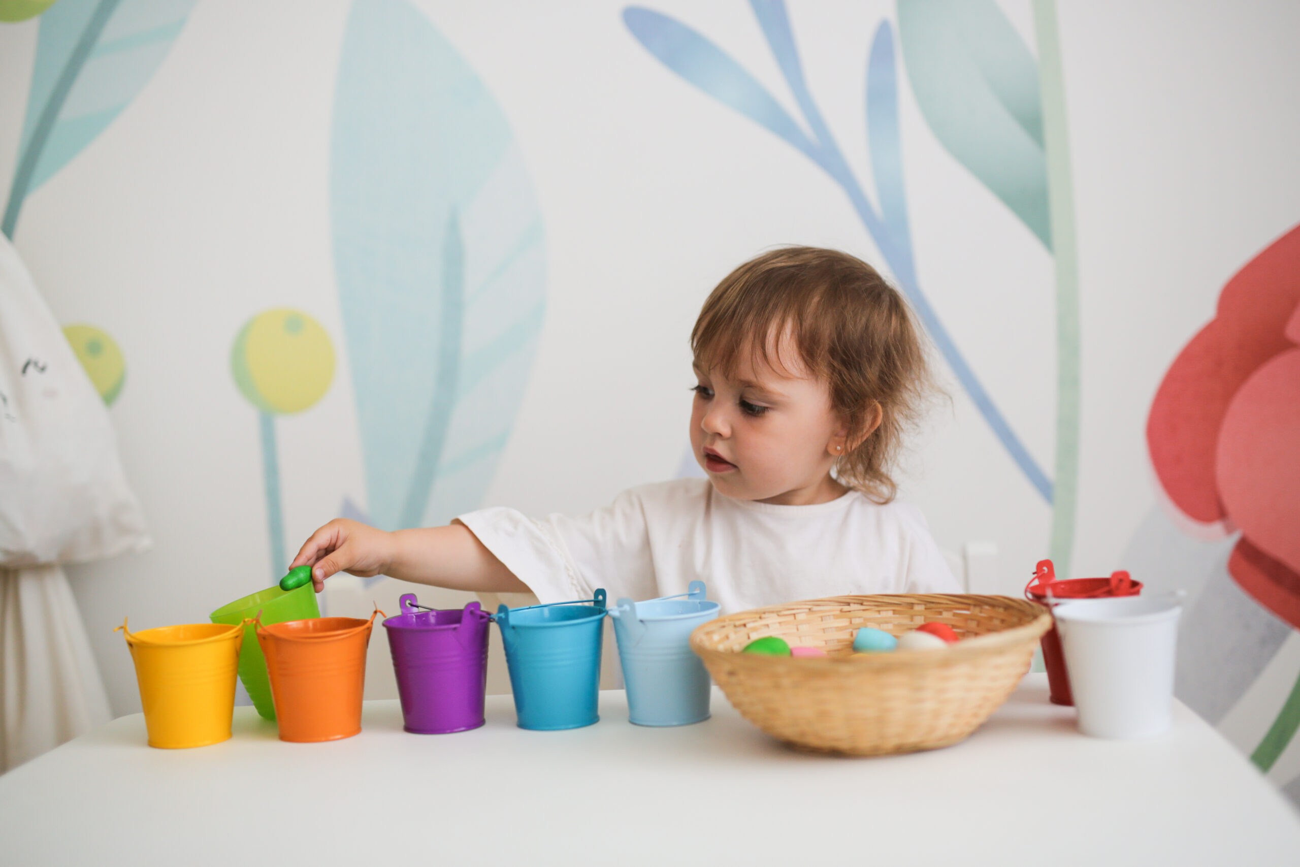 Learning colors, a child plays with colored wooden toys, sorting by color. The concept of earlier child development. A girl with wavy hair is independently engaged at a table in a bright room.